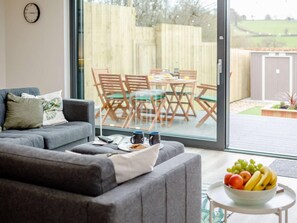 Ein Wohnzimmer mit grauem Sofa, einem Couchtisch mit Obstschale und Blick auf eine Terrassenanlage mit Holztisch und Stühlen.