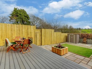 Een houten terras met een tafel en stoelen, een klein tuinpartijtje met gras en een plantenbak, en een houten omheining.
