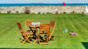 A grassy outdoor area with a wooden table and chairs, a stone wall, and a view of the sea. - 3 Bed in Seahouses (oc-90727) (Seahouses)