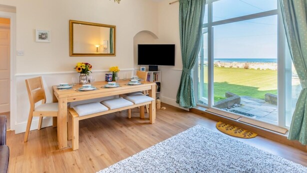 A dining area with a wooden table, chairs, a chandelier, a TV, and a large window with curtains. - 3 Bed in Seahouses (oc-90727) (Seahouses)