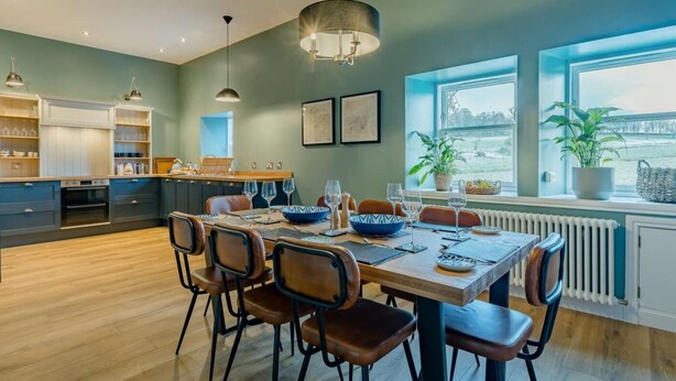 Una cocina moderna con armarios de madera, un comedor con una mesa dispuesta para cuatro personas y ventanas amplias con vistas.
