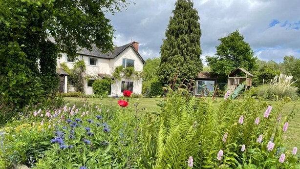 A garden with various plants and flowers, a white house, and a tall tree. - 3 Bed in Malvern (oc-90775) (Mathon)