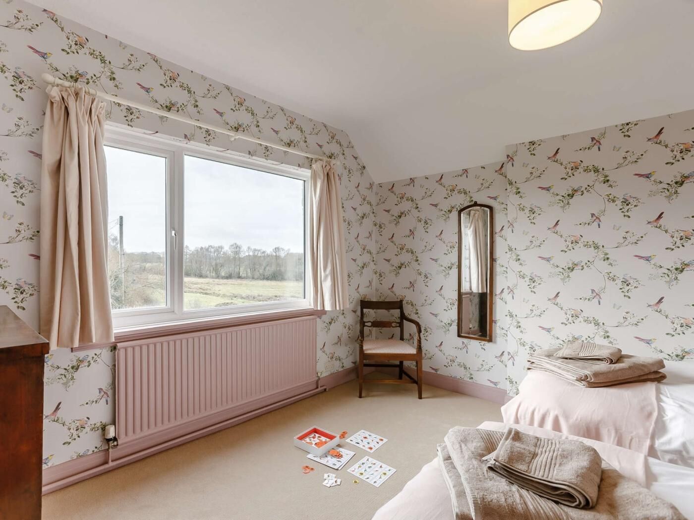 Un dormitorio con ventana, radiador, una silla de madera, espejo y una cama con toallas dobladas.
