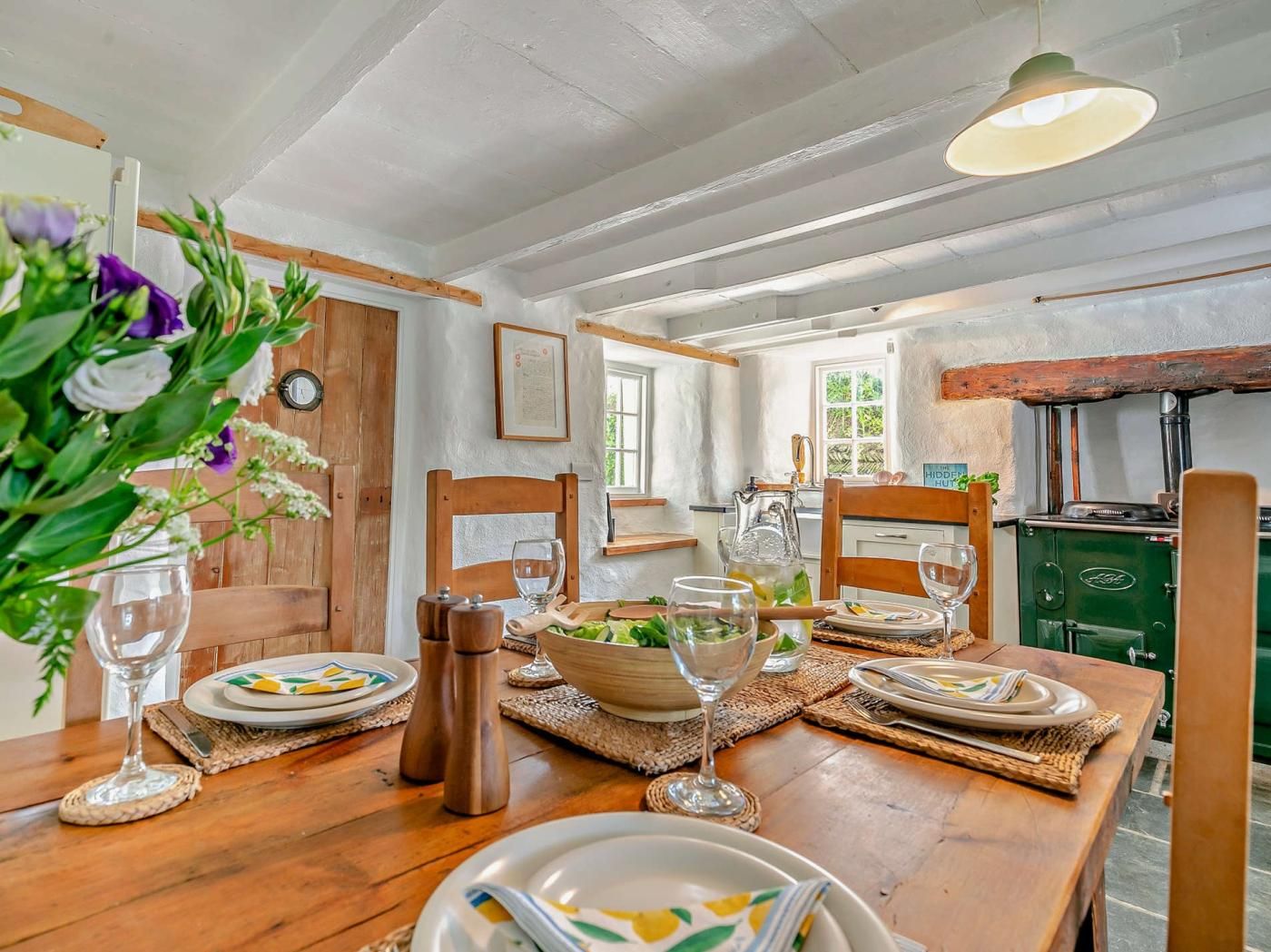 A dining area with a wooden table set for four, surrounded by chairs, with a green stove in the background.