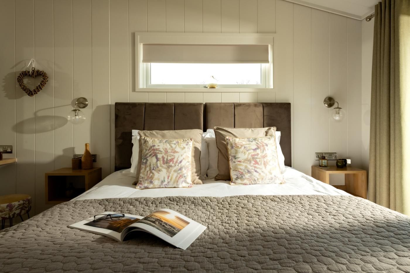A bedroom with a large bed, two pillows, a bedside table with a lamp, a window with a view, and a magazine on the bed.