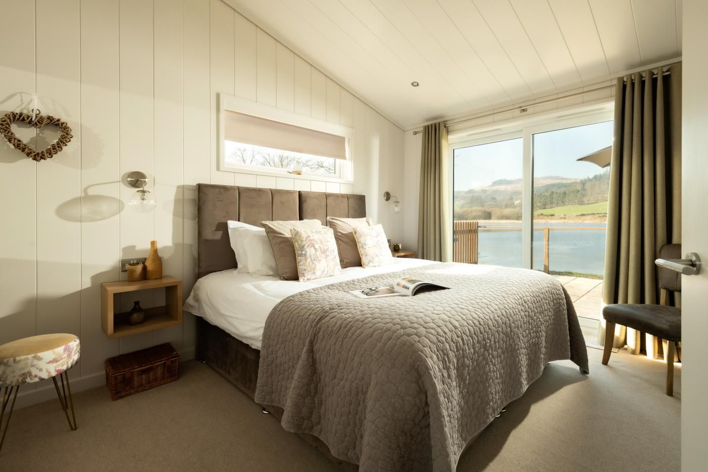 A bedroom with a large bed, a bedside table, a nightstand, a window with a view of the outdoors, and a door.