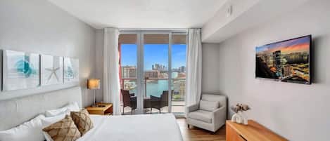 A modern hotel room with a large bed, a TV, a chair, and a view of the city through large windows.
