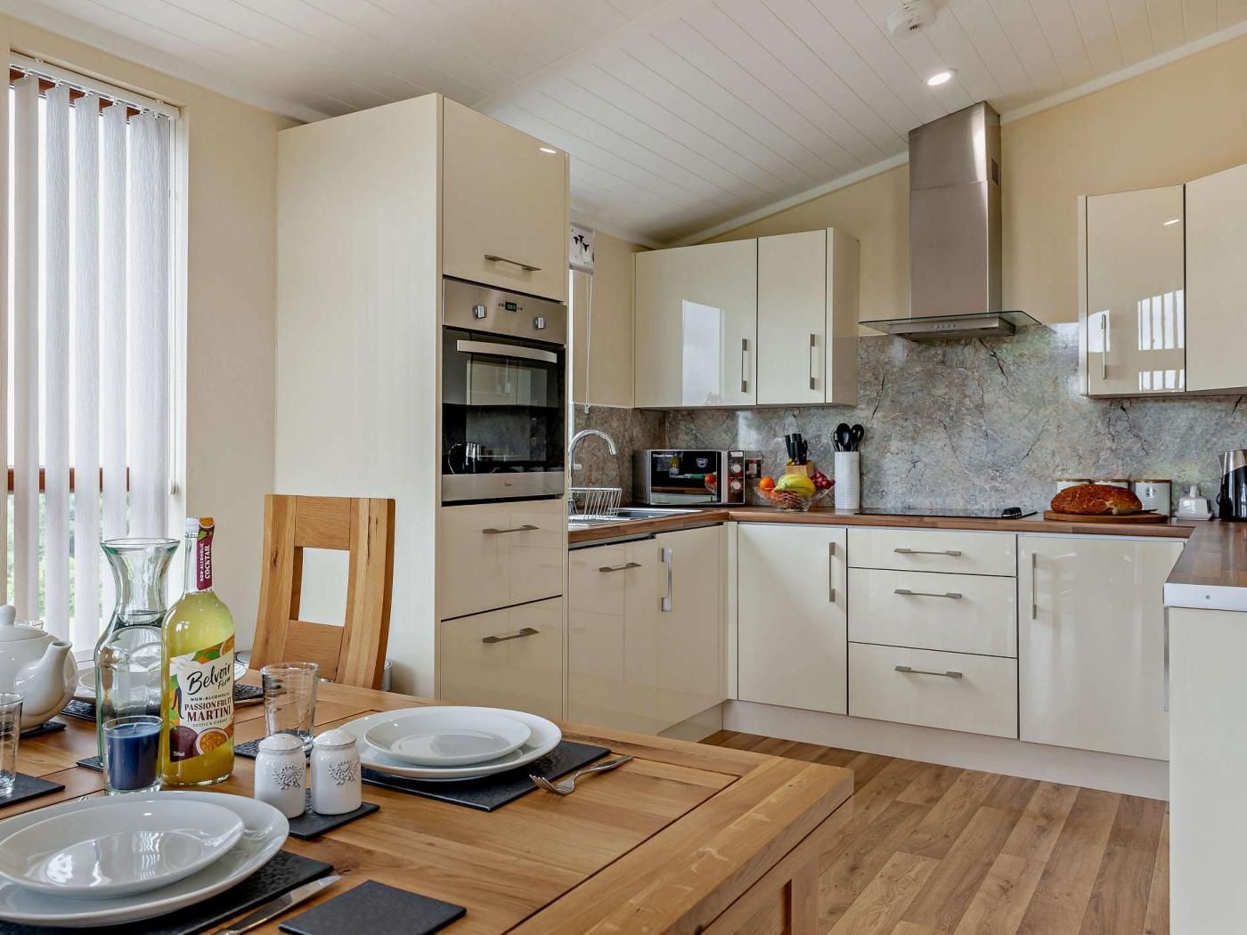 Een moderne keuken met inbouwapparatuur, een eethoek met een houten tafel gedekt voor twee personen, en een raam met gordijnen.