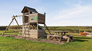 A wooden play structure with a slide and climbing area, surrounded by a picnic table and benches, set in a grassy field.