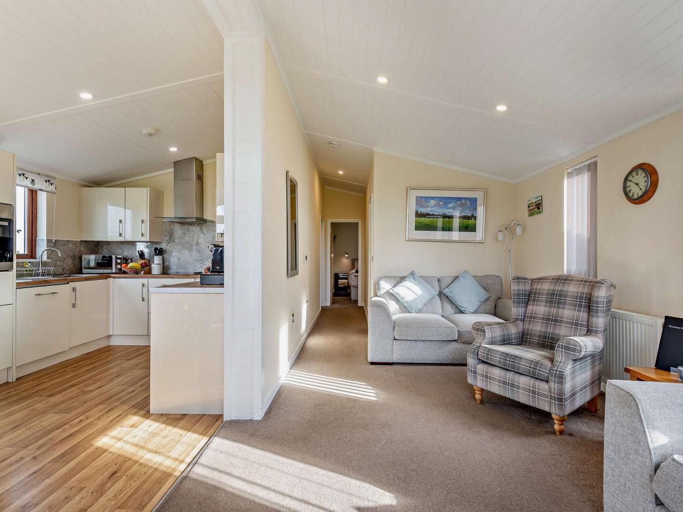A modern kitchen with white cabinets, a central island, and a dining area with a grey sofa and plaid armchair.