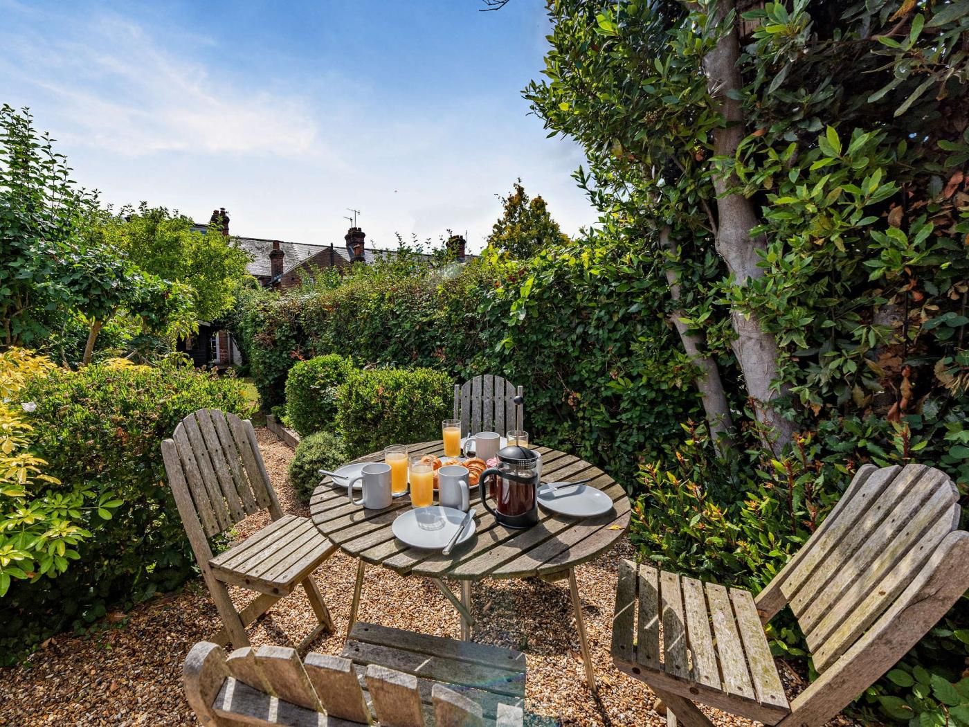 An outdoor wooden table set with plates, glasses, and a pitcher, surrounded by wooden chairs in a garden with dense foliage and a building in the background.