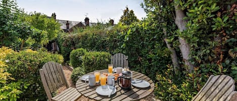 An outdoor wooden table set with plates, glasses, and a pitcher, surrounded by wooden chairs in a garden with dense foliage and a building in the background.