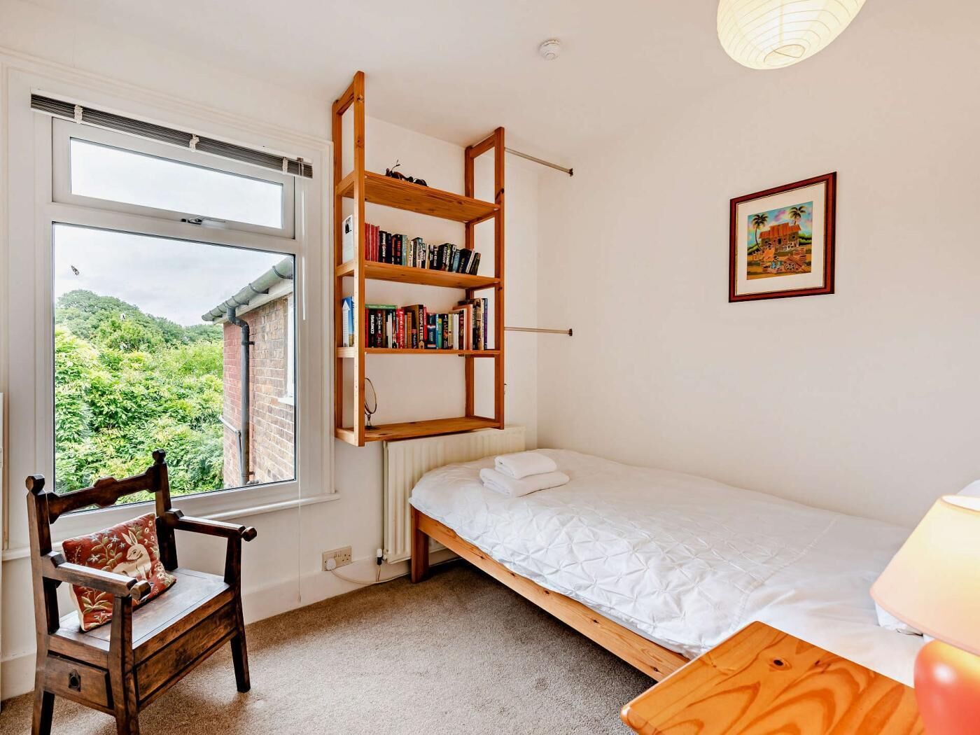 Ein Schlafzimmer mit einem Einzelbett, einem Holzregal, einem Stuhl, einem Fenster mit Blick auf Grünflächen und einem gerahmten Bild an der Wand.