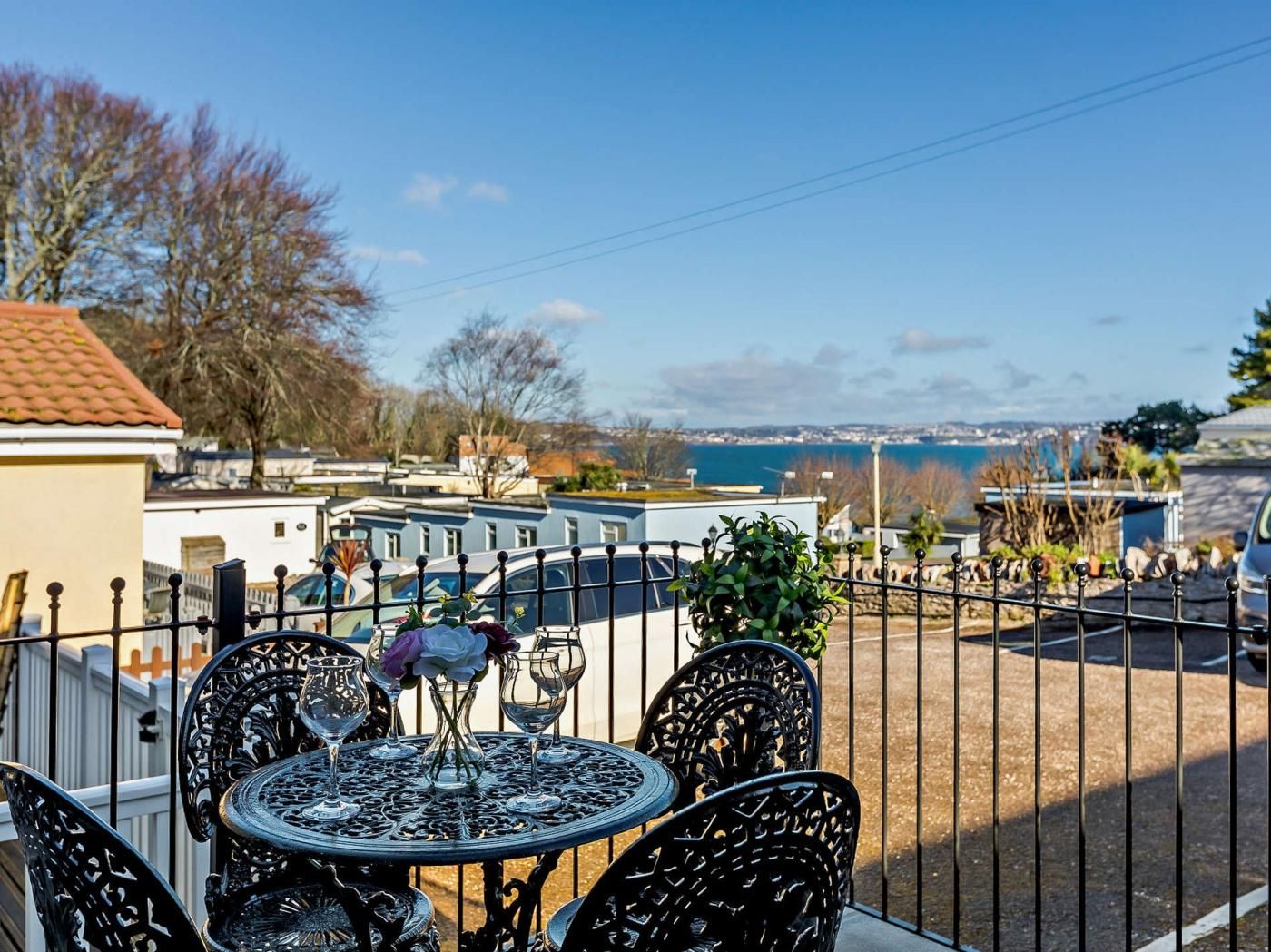 A patio with a table and chairs, overlooking a coastal view with a fence and parked cars.