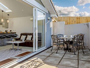 A patio area with a table and chairs, a sliding glass door leading to a living room, and a wooden fence in the background. - 2 Bed in Brixham (oc-90912) (Brixham)