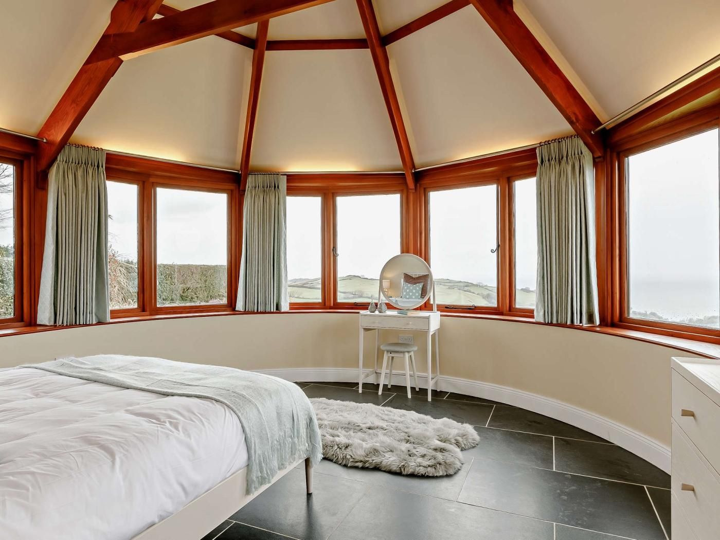 A bedroom with a bed, a desk with a mirror, and large windows with curtains.