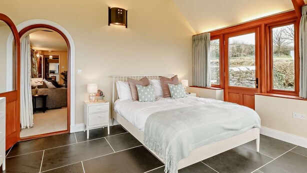Un dormitorio con cama, mesitas de noche, lámpara, ventana con cortinas y vistas al exterior.
