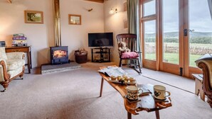 Una sala de estar acogedora con chimenea, televisor, un osito de peluche y vistas al campo a través de amplios ventanales.