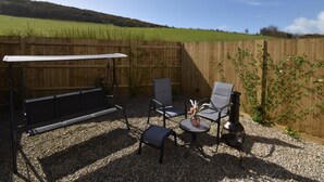Eine Terrasse mit Tisch und Stühlen, einem Grill und Blick auf ein grünes Feld und eine Hügelkuppe.