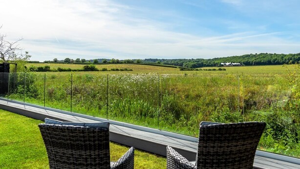 A view from a balcony with wicker chairs, overlooking a grassy field and a distant white building. - 4 Bed in Holt (oc-91228) (Weybourne)