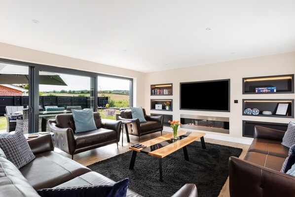 A modern living room with leather sofas, a flat-screen TV, and a fireplace. - 4 Bed in Holt (oc-91228) (Weybourne)