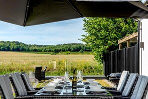 An outdoor dining area with a table set for four, surrounded by chairs, and a view of a field and trees in the background. - 4 Bed in Holt (oc-91228) (Weybourne)