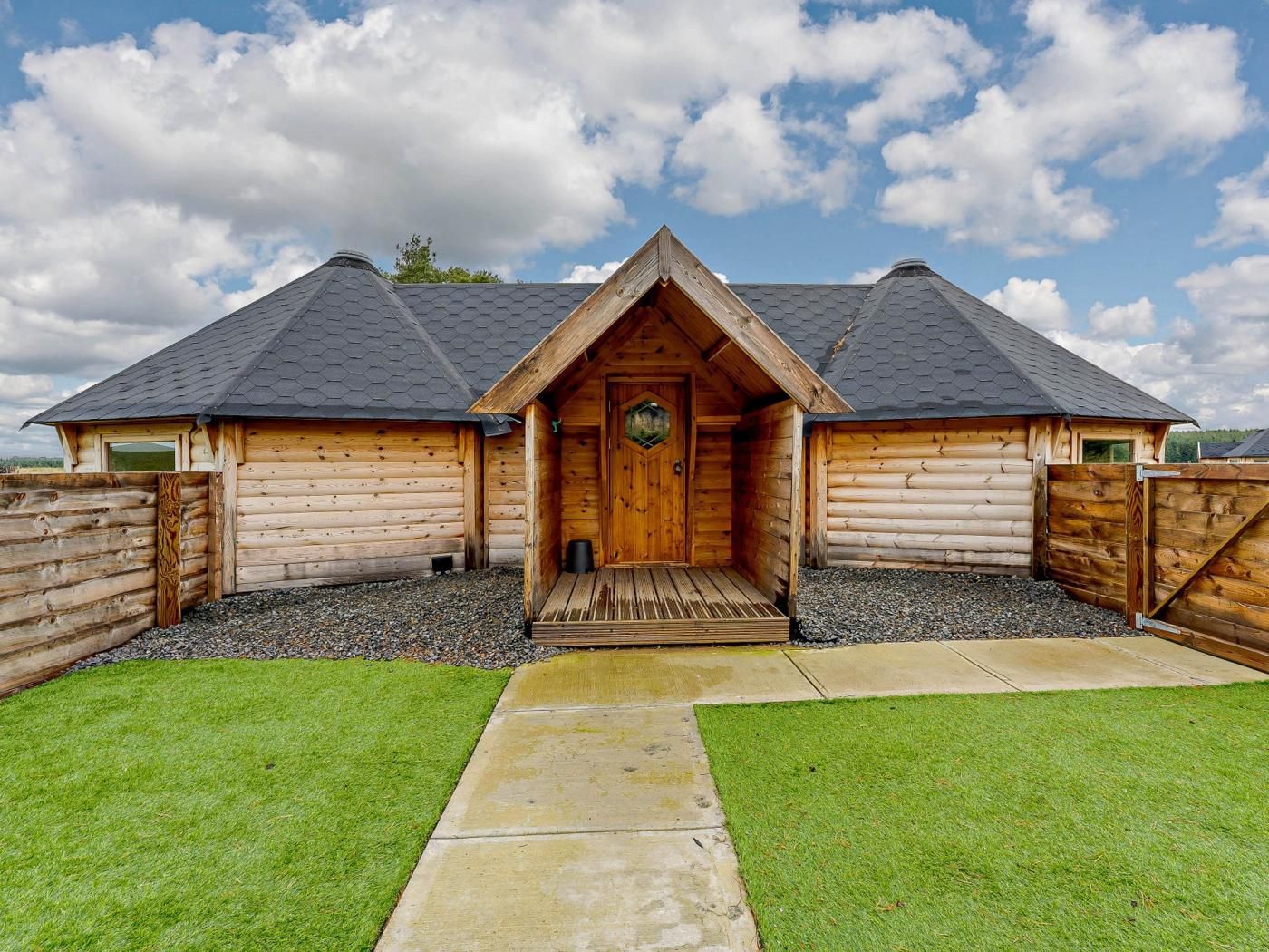 A log cabin with a central entrance, a gravel driveway, and a grassy lawn.