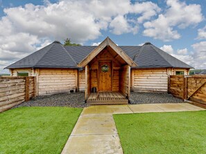 Une cabane en rondins avec une entrée centrale, une allée en gravier et une pelouse verdoyante.