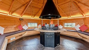 A wooden cabin interior with a central fireplace, seating areas, and a herringbone floor pattern.