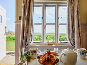 A breakfast setup with a view of a green landscape through a window, a table with a teapot, cups, and pastries. - 1 Bed in Camelford (oc-90818) (Forrabury and Minster)