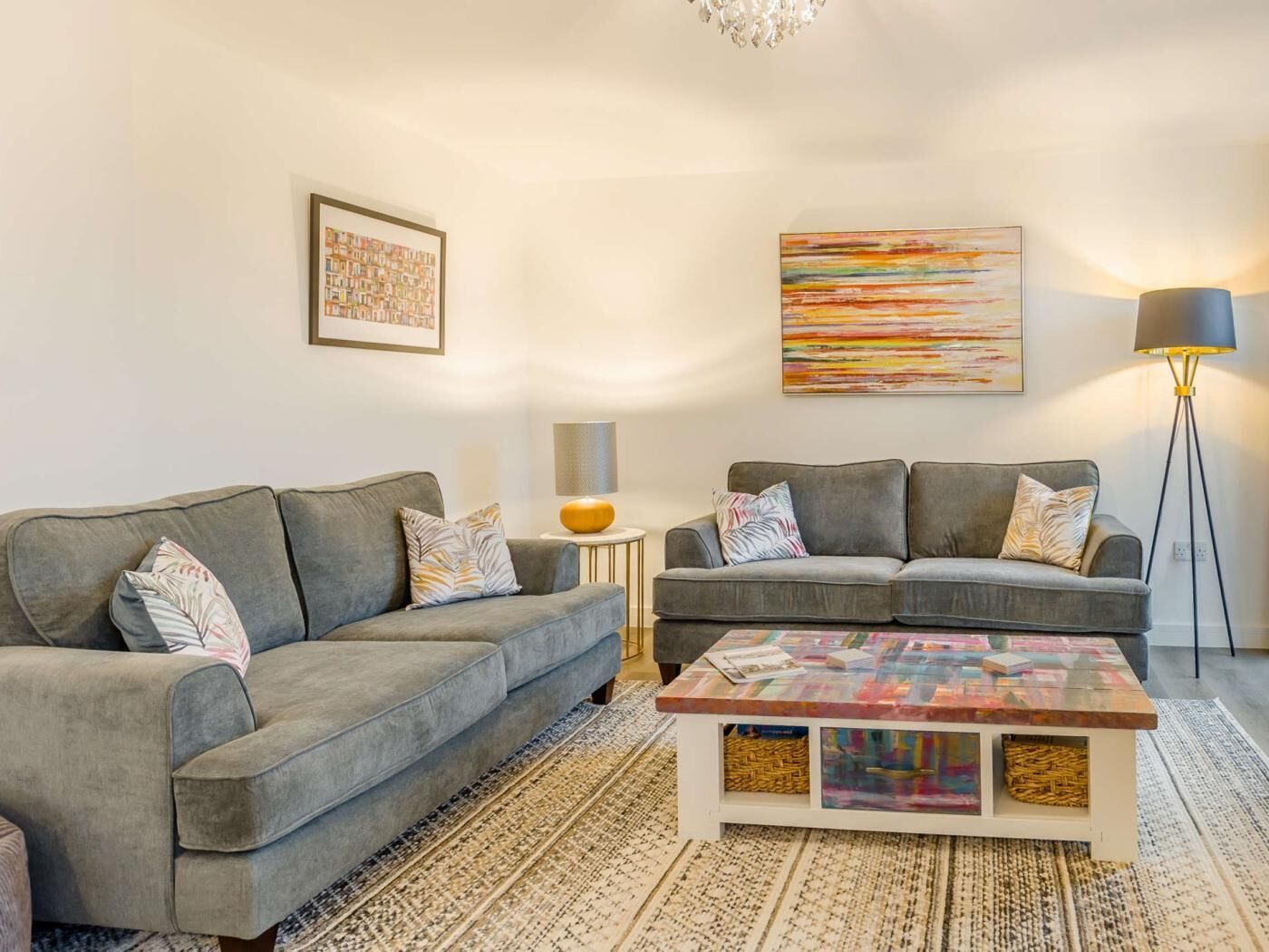 A modern living room with two grey sofas, a coffee table with a colorful top, a floor lamp, and two wall art pieces.