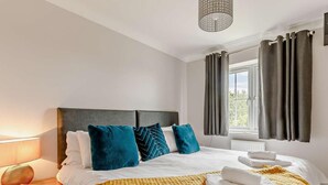 Ein Hotelzimmer mit einem Bett, einem Nachttisch, einer Lampe, einem Fenster mit Vorhängen und Blick ins Grüne.