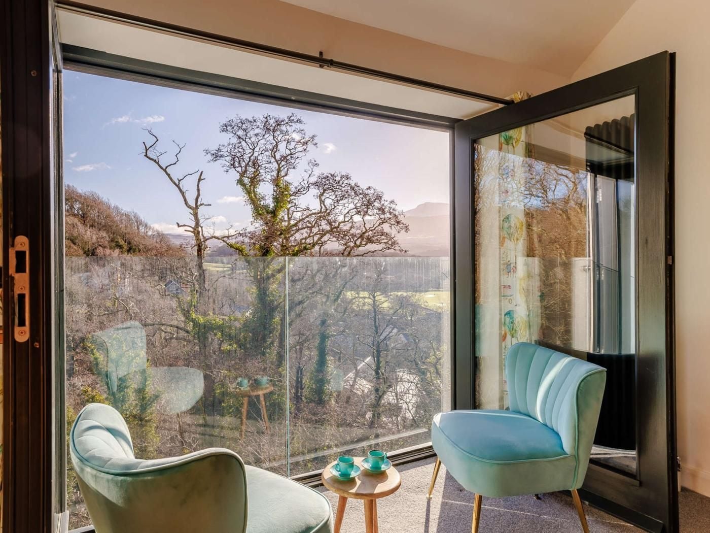 Une chambre moderne dotée d’une grande porte vitrée donnant sur un paysage extérieur magnifique, de deux fauteuils rembourrés, d’une petite table en bois sur laquelle se trouvent une tasse et une soucoupe, et d’une cheminée encadrée de noir.