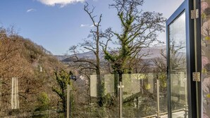 Un balcó amb barana de vidre, vista a arbres i turons, i cel serè.