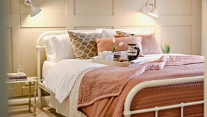 Een net opgemaakt bed met een roze deken, een nachtkastje met een glas water, en een dienblad met een koffiezetapparaat en een boek.