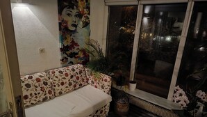 Huoneessa on kukkapainokuosinen sohva, suuri ikkuna, josta on näkymä, patteri ja seinällä oleva taulu.