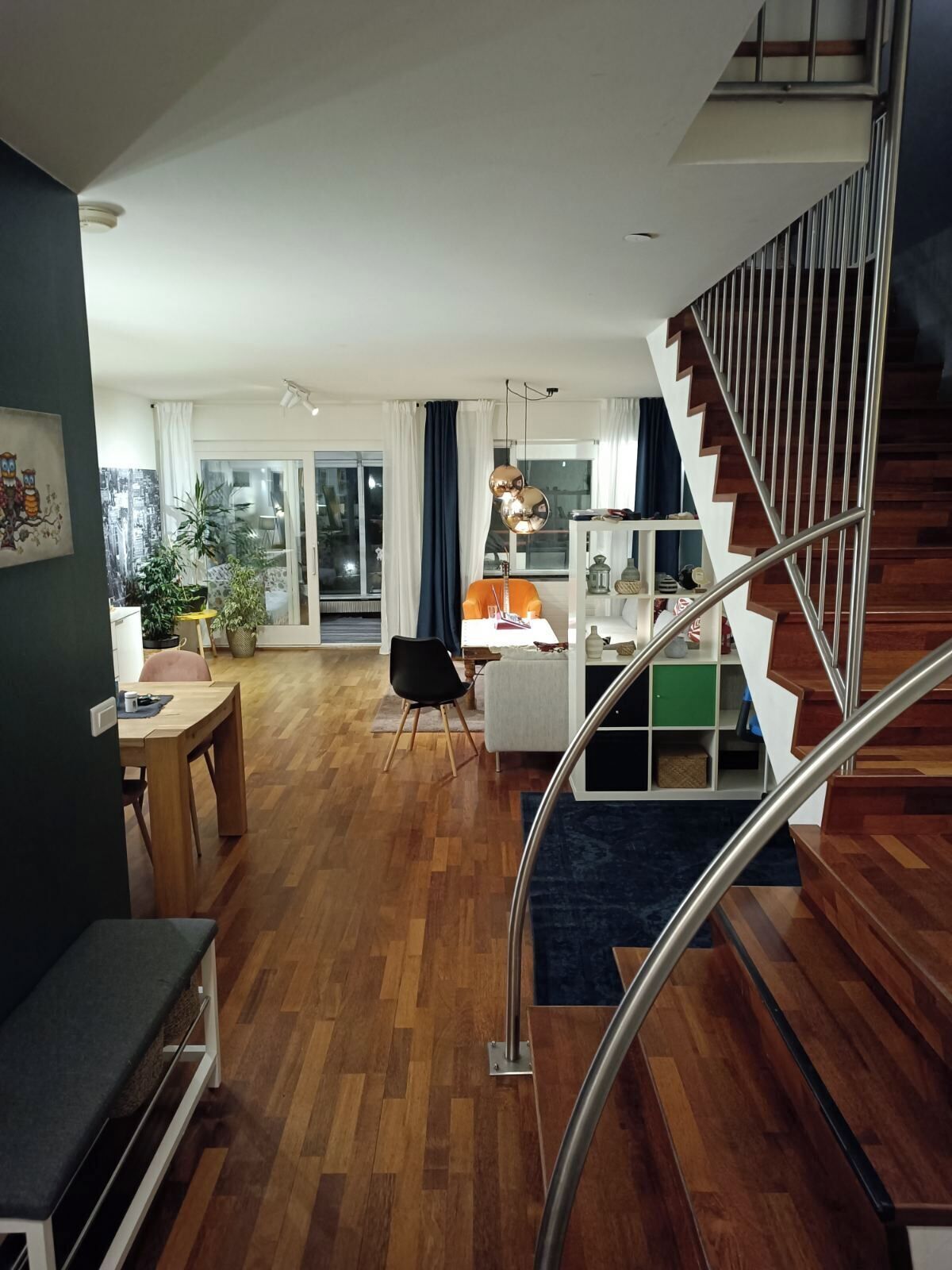 A modern living room with a wooden floor, a staircase with metal railings, a dining area with a table and chairs, and a view of the outside through a glass door.