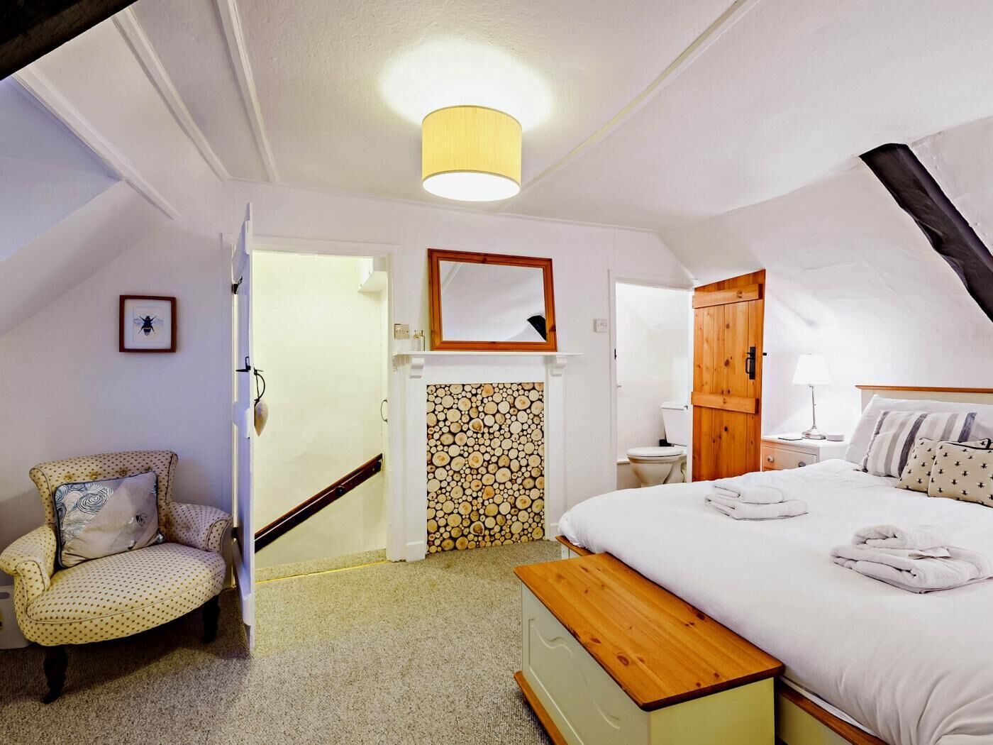 Ein Schlafzimmer mit einem Bett, einem Sessel, einer hölzernen Bank, einem Spiegel, einer Toilette und einer Tür zu einem anderen Raum.