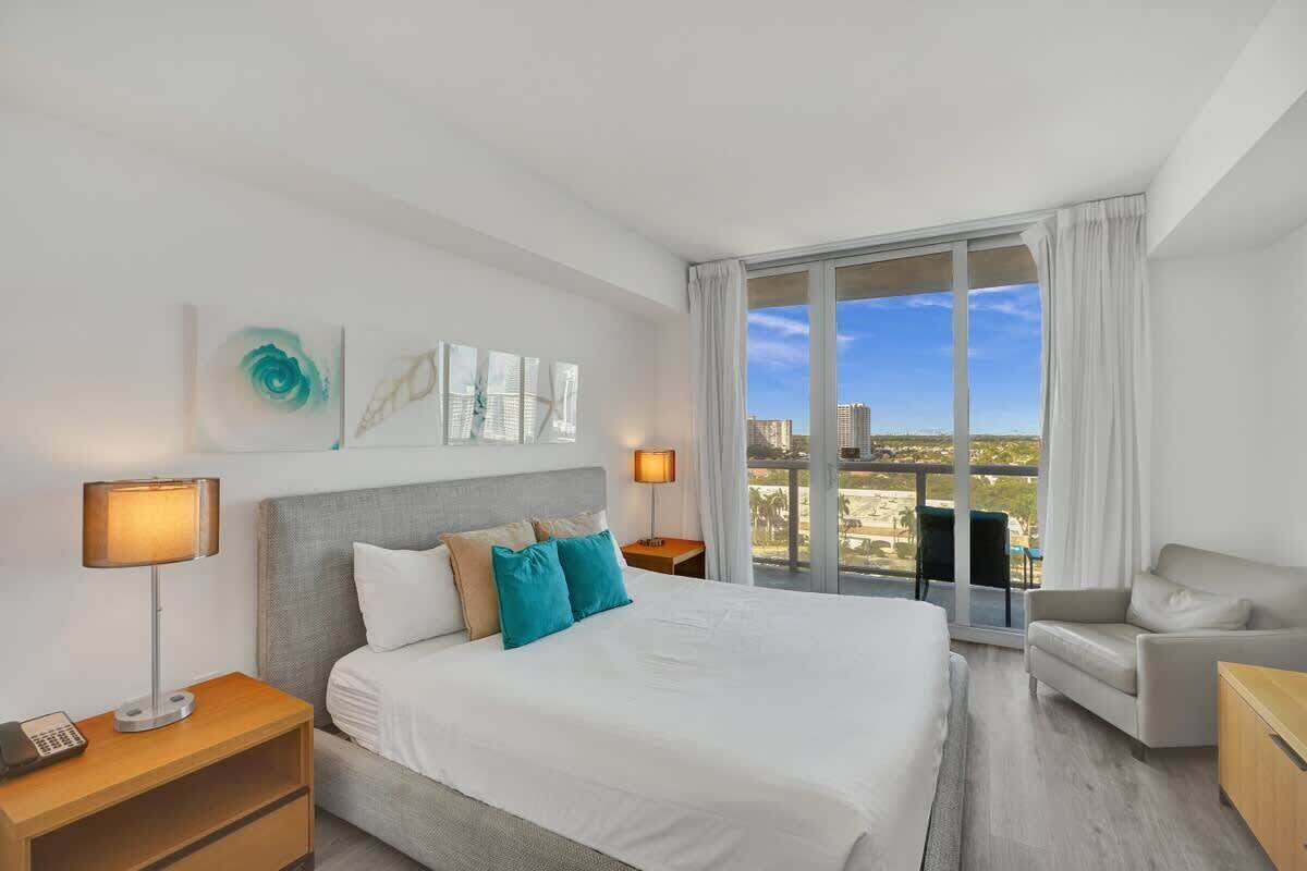 Une chambre d’hôtel avec un grand lit, deux tables de chevet, une commode, un canapé et une vue sur la ville grâce à de grandes fenêtres.