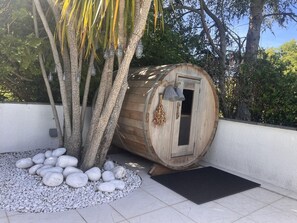 Eine Holztonne als Sauna mit Tür, umgeben von weißen Kieselsteinen und einer schwarzen Matte, mit Bäumen und einer weißen Wand im Hintergrund.