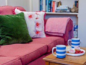 A cozy living area with a sofa, pillows, a coffee table with a plate of pastries, and a bookshelf in the background. - 3 Bed in Bideford (oc-90864) (Weare Giffard)