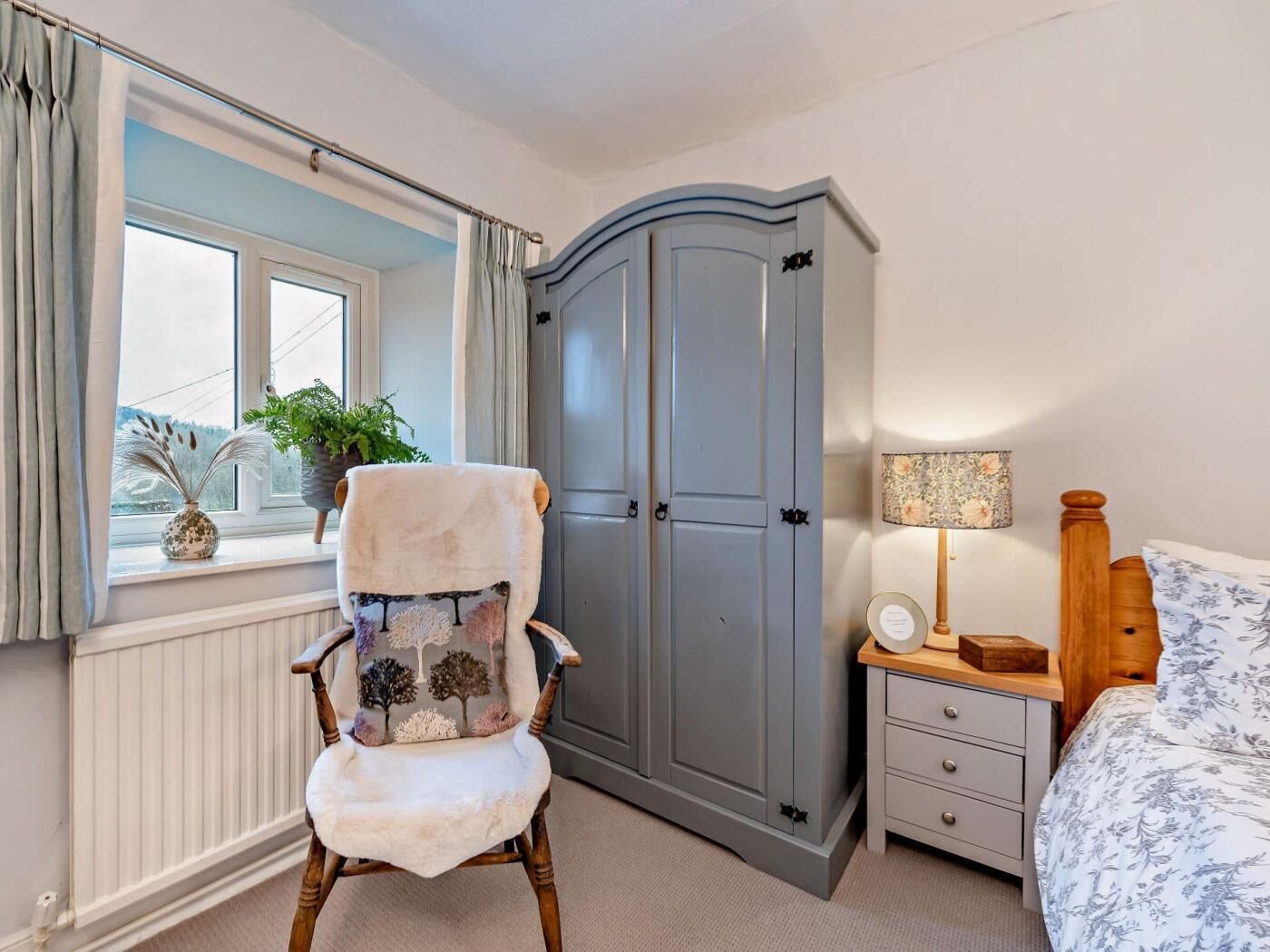 Une chambre avec une chaise en bois, une armoire grise, une table de chevet avec une lampe et une fenêtre avec des rideaux.