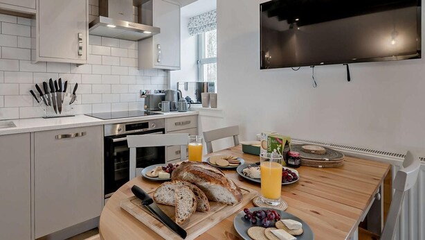 A modern kitchen with white cabinets, a wooden dining table, and a flat-screen TV mounted on the wall. - 3 Bed in Crieff (oc-90794) (Comrie)