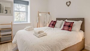 A neatly made bed with white bedding and red and white pillows, a wooden side table, a window with a view of trees, and a heart-shaped decoration on the wall. - 3 Bed in Crieff (oc-90794) (Comrie)