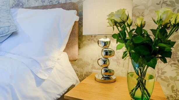 Un lit bien fait, avec du linge de lit blanc, une lampe de chevet dotée d’un abat-jour réfléchissant et un vase contenant des plantes vertes, posé sur une table de chevet en bois.