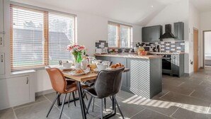 A modern kitchen with a dining area, featuring a central island, grey cabinets, and a large window with blinds. - 2 Bed in King's Lynn (oc-91275) (North Runcton)