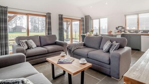 A modern living room with two grey sofas, a wooden coffee table, and a kitchen area with large windows. - 2 Bed in King's Lynn (oc-91275) (North Runcton)