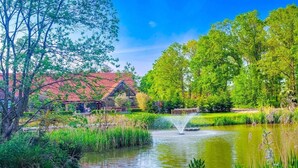 A serene pond with a fountain, surrounded by lush greenery and a traditional building with a red roof. - 2 Bed in King's Lynn (oc-91275) (North Runcton)