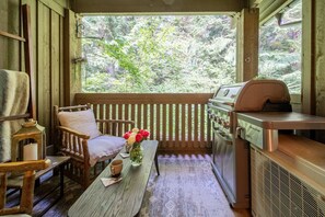 Eine Holzterrasse mit Grill, einem gepolsterten Stuhl, einem Tisch mit Kerze und Blumen und Blick auf einen Wald.