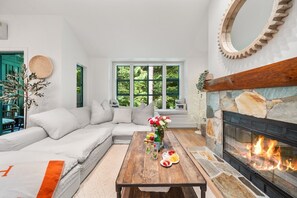 A living room with a sectional sofa, a fireplace, and a large window with a view of greenery. - Near Gondola & Lake | Private Hot Tub| Free Ski Shuttle & Parking (Whistler)